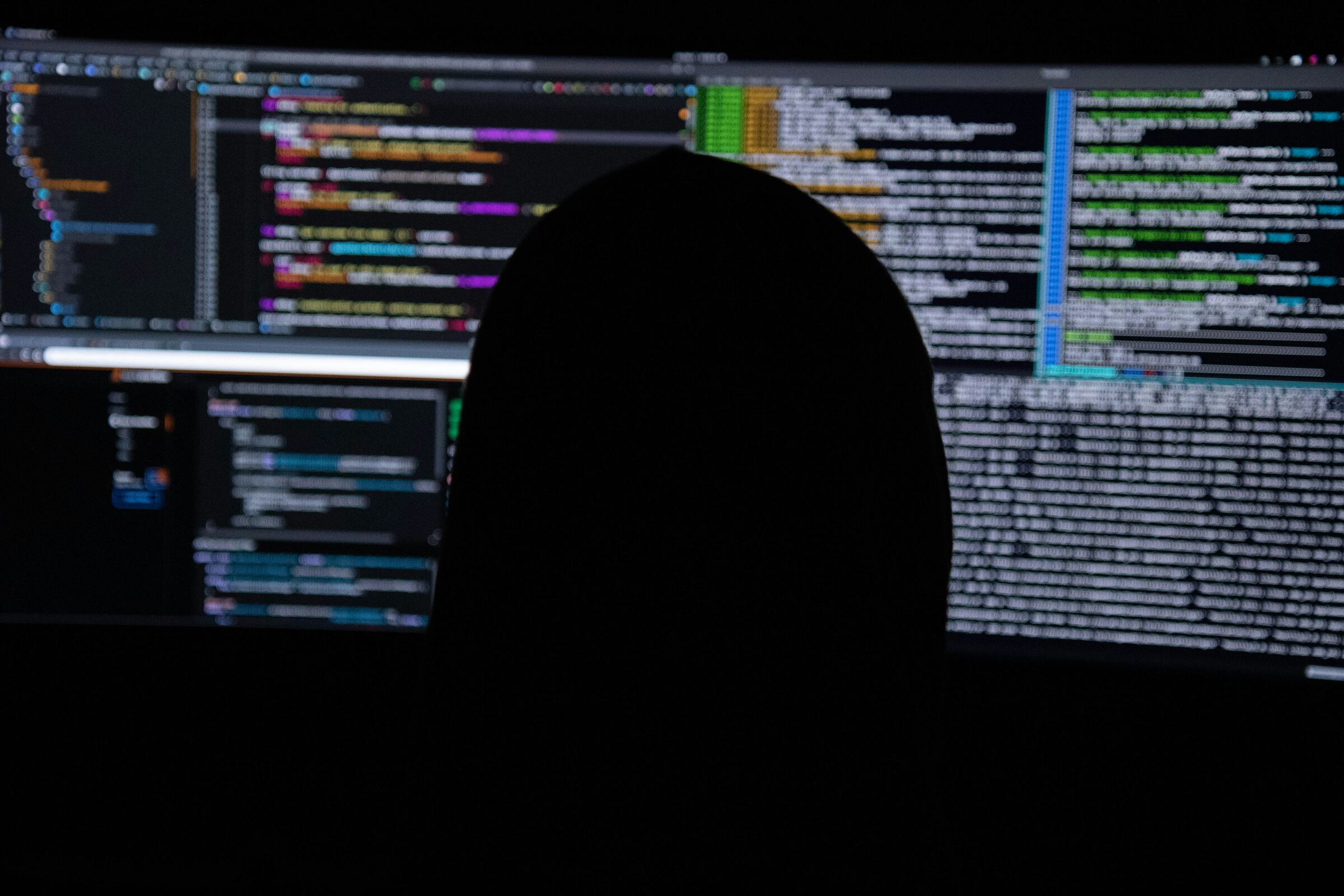 Hooded figure in front of monitors showing code